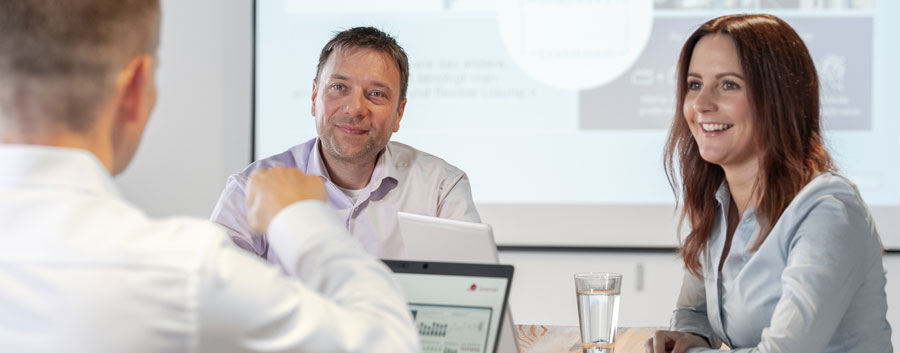 Das Bild zeigt einen Vertriebsmitarbeiter und eine Vertriebsmitarbeiterin, die mit Laptops an einem großen Tisch sitzen. Sie hören einer Person zu, die im Vordergrund des Bilds zu sehen ist.